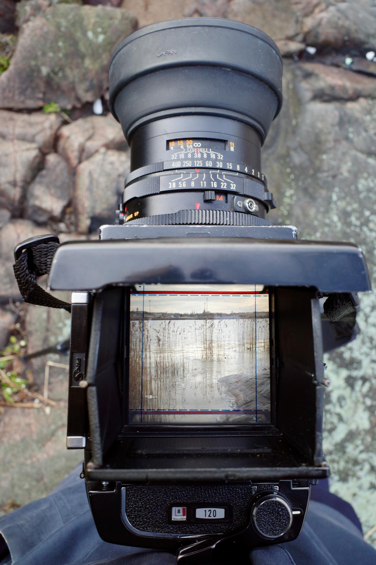 The medium-format camera seen from the above. The viewfinder shows a landscape with rocky seashore and reeds in the front and a city in the background.