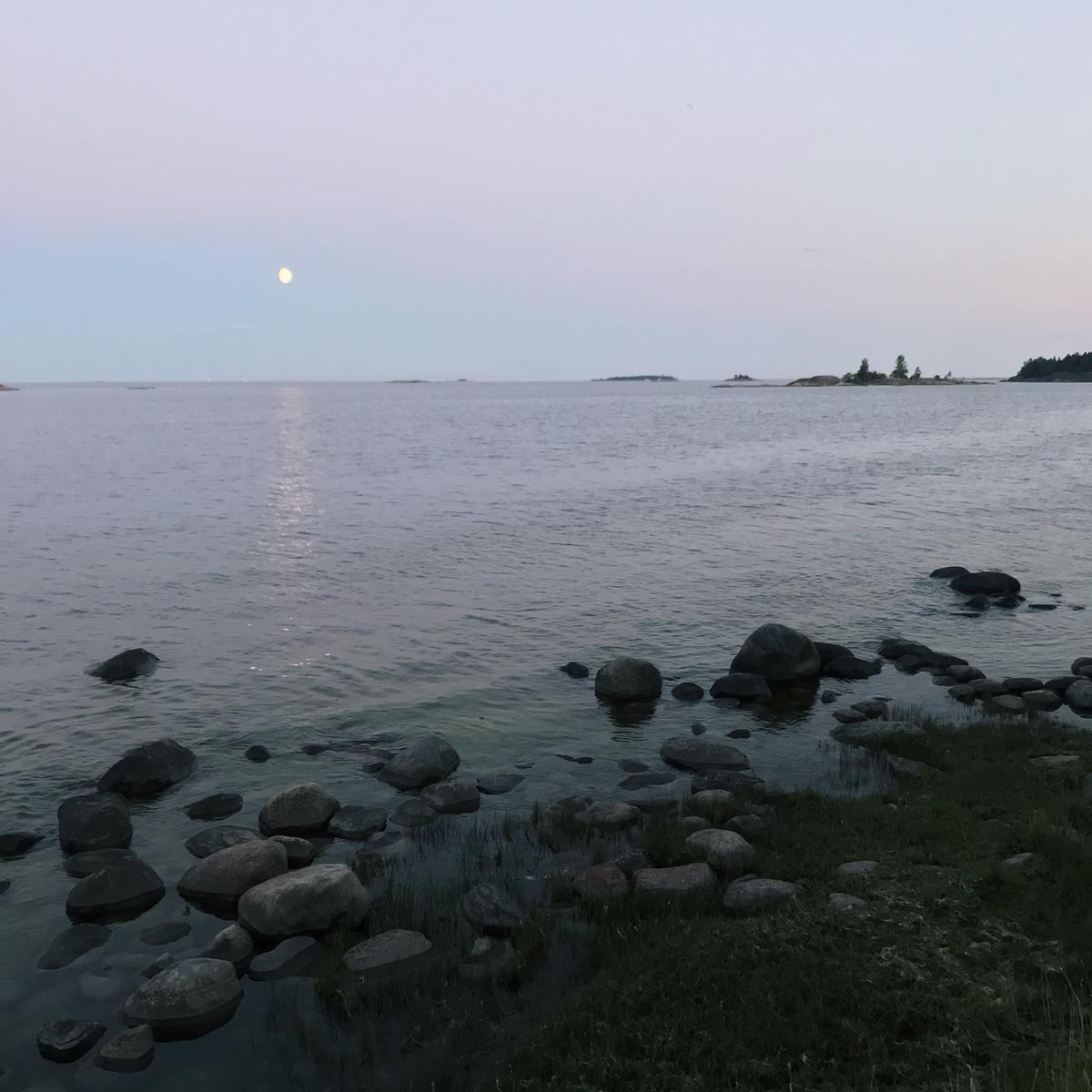 Moon shining over the sea in the evening. There's a rocky shore close to the camera and small islands in the horizon.