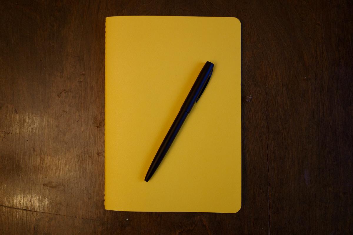 A yellow notebook laying on a table with a black ballpoint pen on top of it.