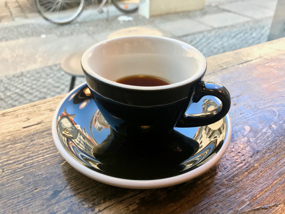 A cup of black filter coffee on a wooden table in front of a window to a street.