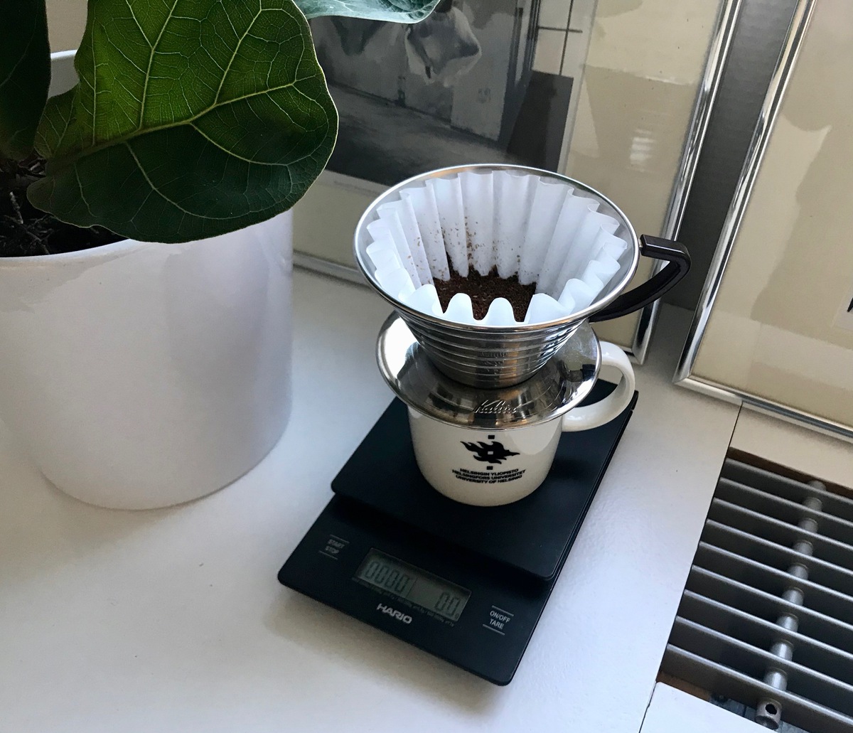 A Kalita filter cone filled with coffee grounds. The cone is on a mug and the mug is on a balance.