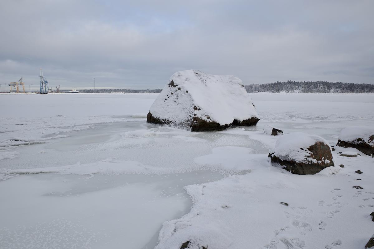 Large snow-covered rock sitting in the frozen sea near shore.
