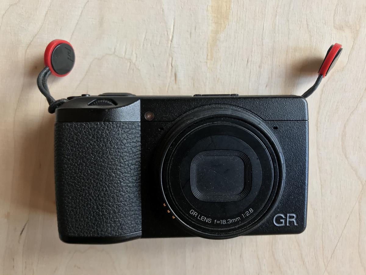 Ricoh GR III, a pocket camera, lying on a plywood surface, lens facing to the viewer. The camera is a bit dusty and the ring cap is missing, exposing a metal connector around the lens.