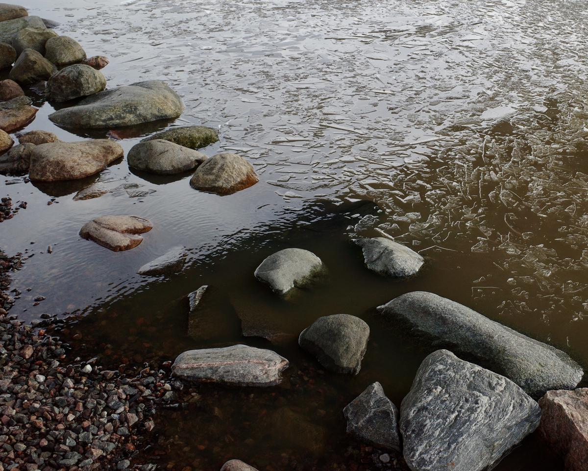 A composition of rocks in the shore. There's a thin, broken layer of ice in the sea.